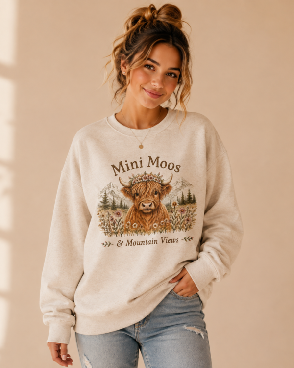 Woman wearing the Mini Moos & Mountain Views Crewneck Sweatshirt featuring a Highland cow, wildflowers, and mountain scenery.