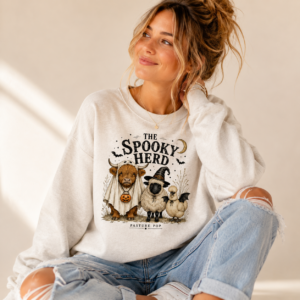 Woman wearing The Spooky Herd Crewneck Sweatshirt featuring a Highland cow, sheep, and Silkie chicken in Halloween costumes.