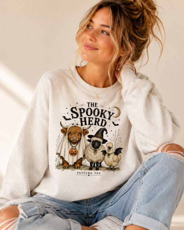 Woman wearing The Spooky Herd Crewneck Sweatshirt featuring a Highland cow, sheep, and Silkie chicken in Halloween costumes.